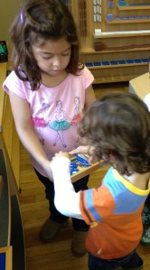 girl helps younger student by getting him some materials to work with and providing basic instructions